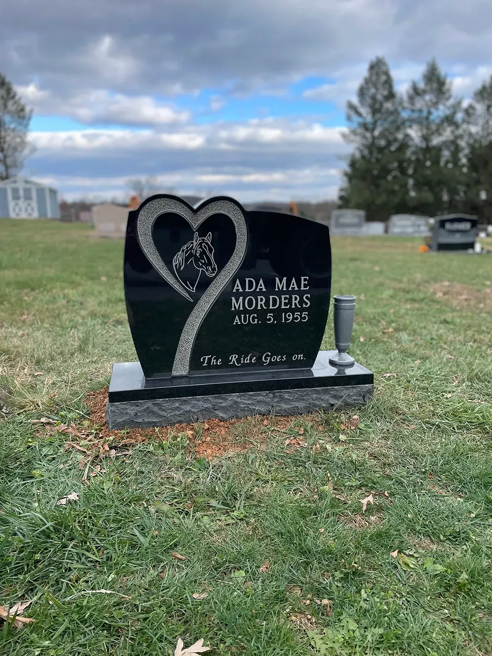 Black granite gravestone featuring a heart design with an engraved horse, set in a grassy cemetery under a cloudy sky.