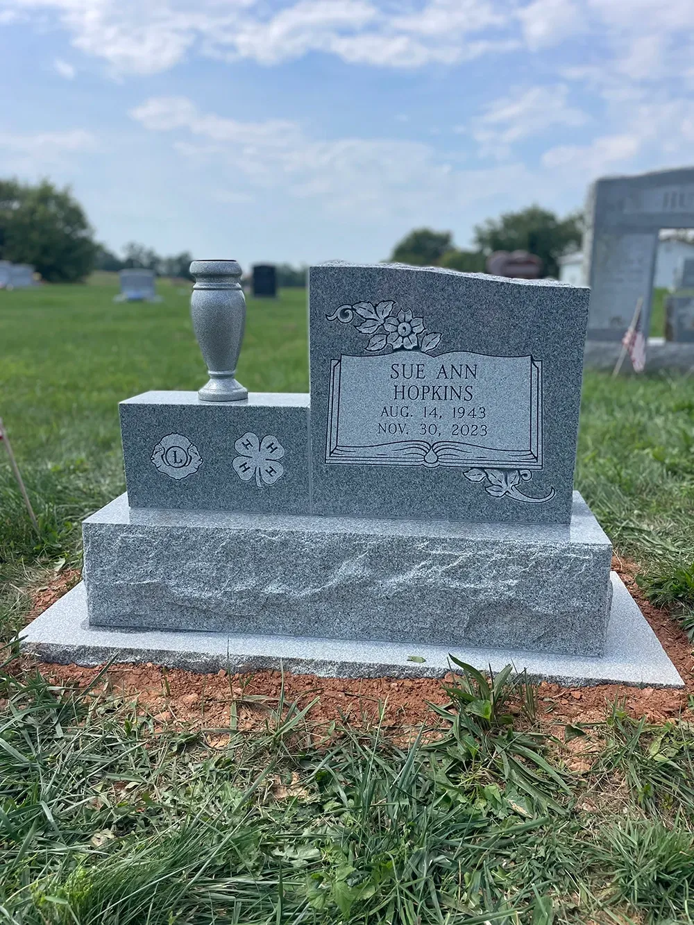 A gray granite headstone in a cemetery with a 4-H emblem and a small vase, marking the grave of Sue Ann Hopkins.