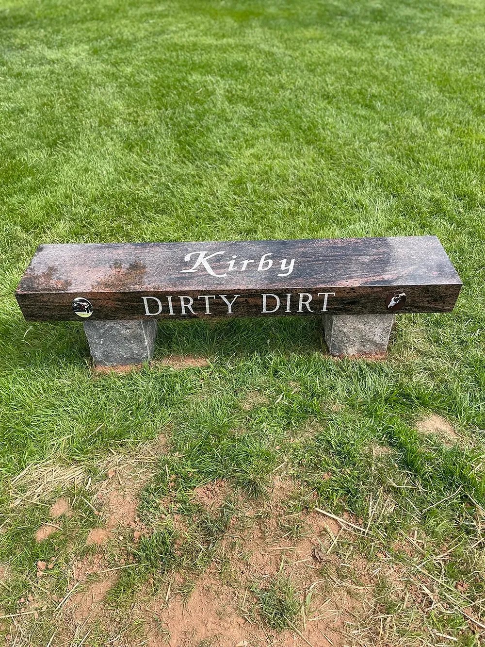 A dark stone bench sitting in a green grassy field with the words 