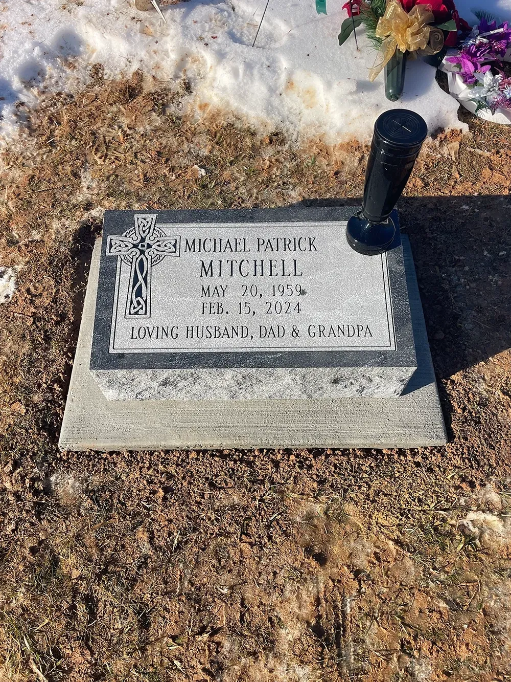 A grey granite grave marker for Michael Patrick Mitchell, featuring an etched Celtic cross and a black vase, in a snowy lot.
