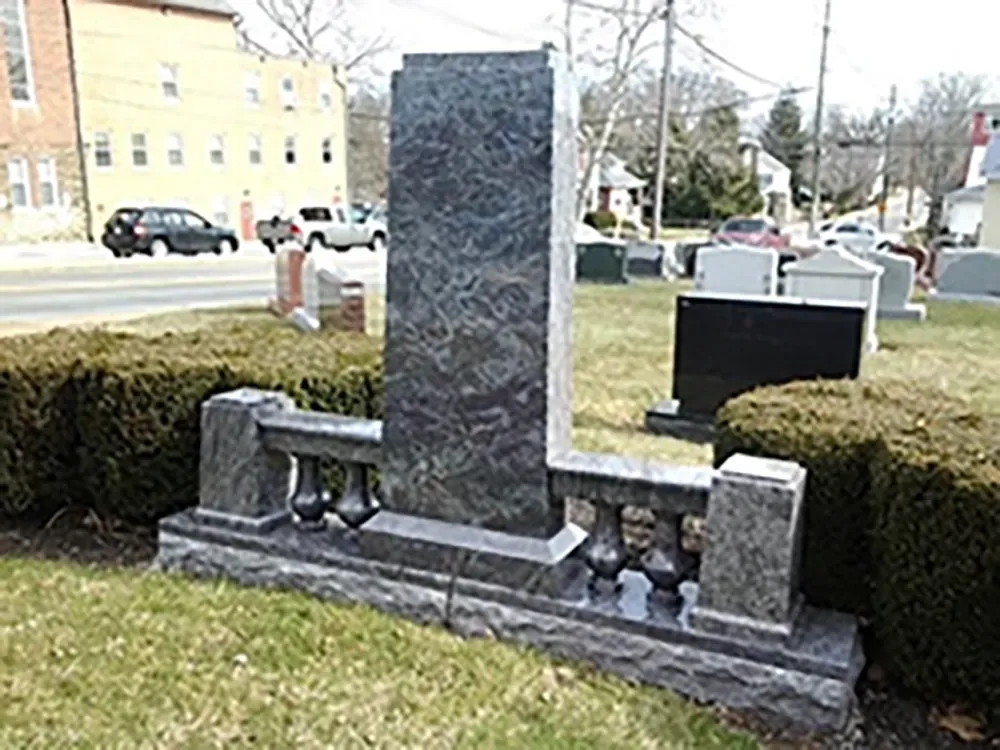 A tall, dark gray granite monument with a balustrade design set in a cemetery alongside a street with parked cars.