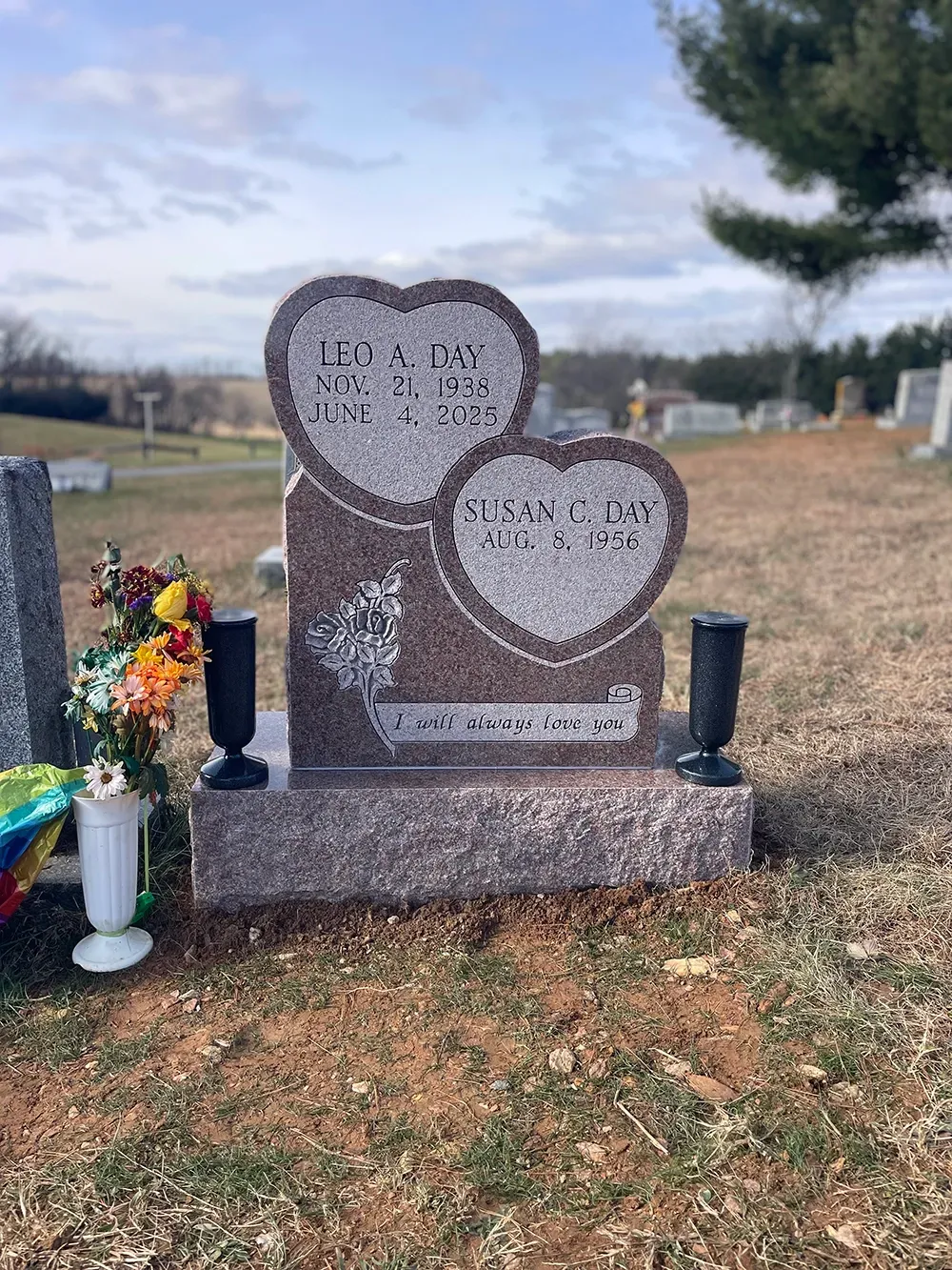 A granite headstone with two heart-shaped engravings, resting in a cemetery with a bouquet of flowers in a white vase.