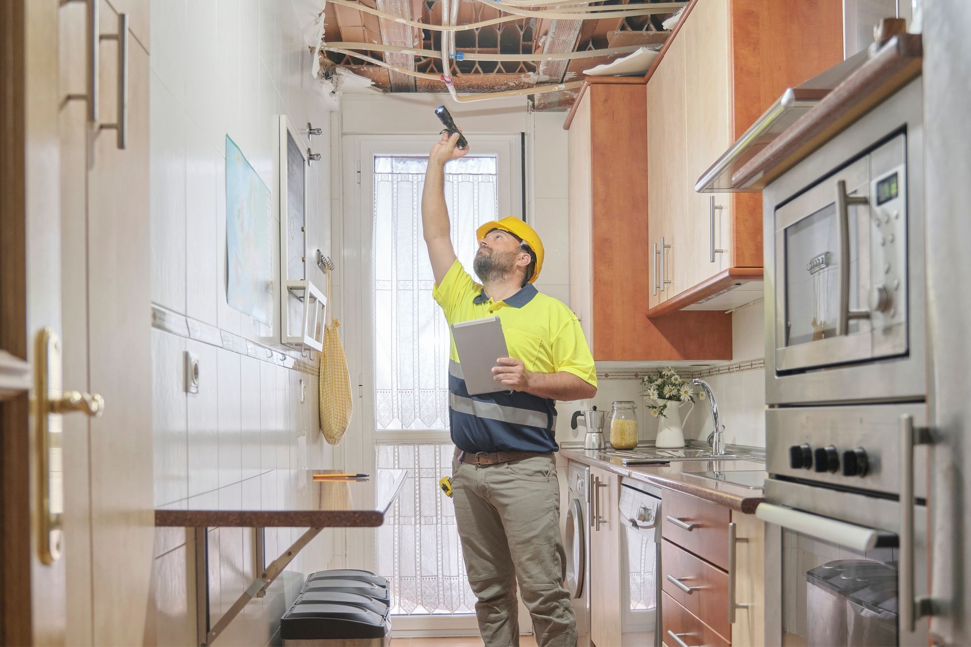 Worker in a yellow shirt inspects water damage on a kitchen ceiling with a flashlight and clipboard.