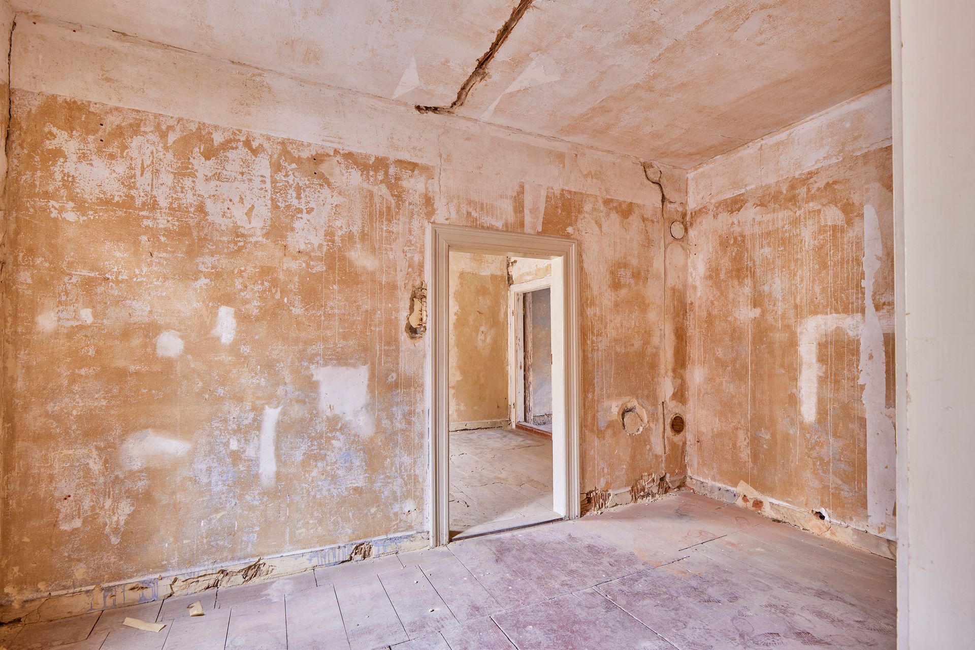 An empty room with cracked walls, peeling paint, and a doorway to another space showing water damage