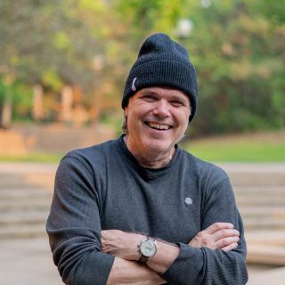 Man in black beanie and gray sweater smiles with arms crossed outdoors.