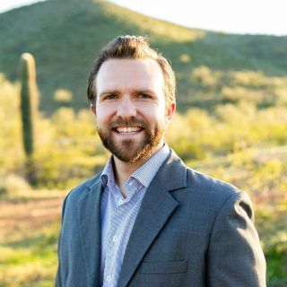 Man in a gray suit smiles, standing outdoors in front of a desert landscape with saguaro cactus.