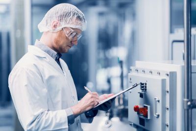 Person in lab coat and hairnet inspecting machinery, taking notes on a clipboard in an industrial setting.