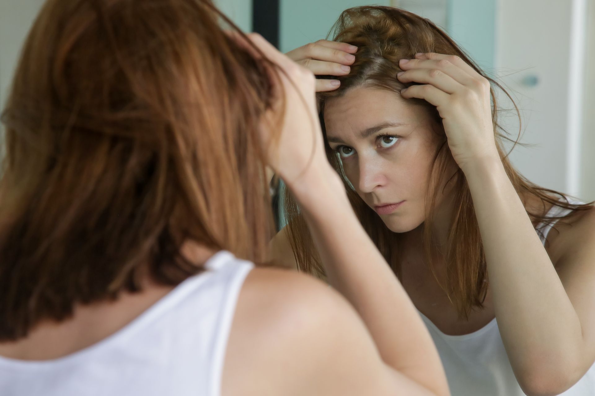 A person looks into a mirror, using their hands to part their hair and examine their scalp.