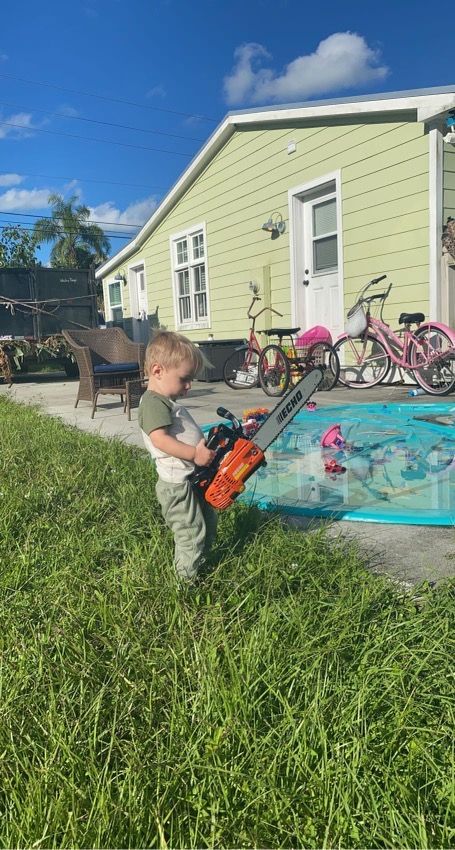 Child holding an orange toy by a backyard kiddie pool beside a light green house and bikes.