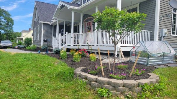 A gray house with a white wrap-around porch features a front garden bed with a small tree and a raised, metal cold frame.