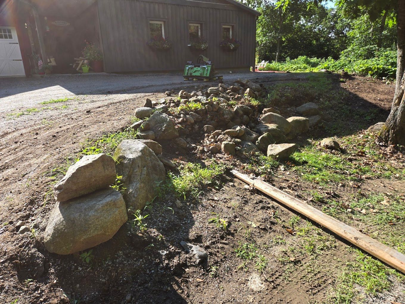 A pile of rocks forming a low garden wall in a yard, with a dark wooden barn and trees in the background.