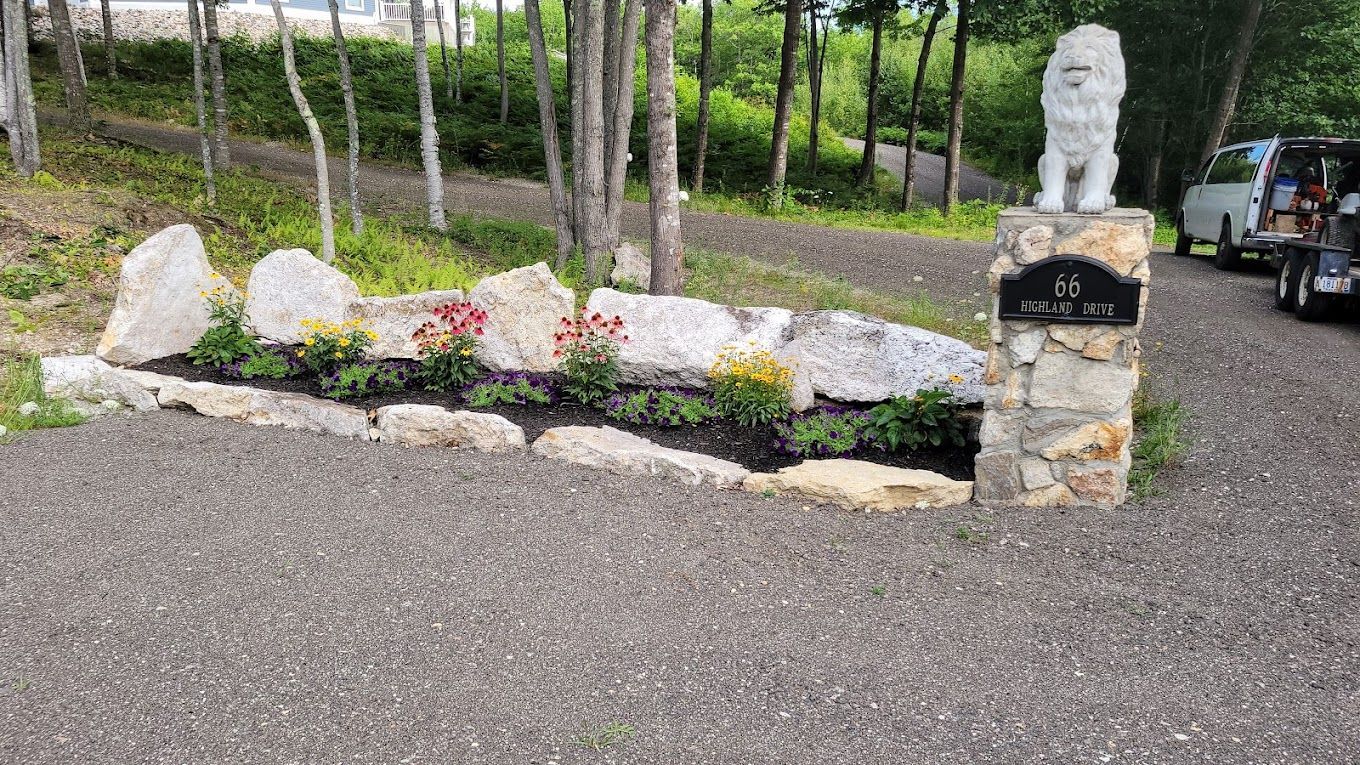 A garden bed lined with light gray boulders, featuring flowers and a stone pillar topped with a lion statue.