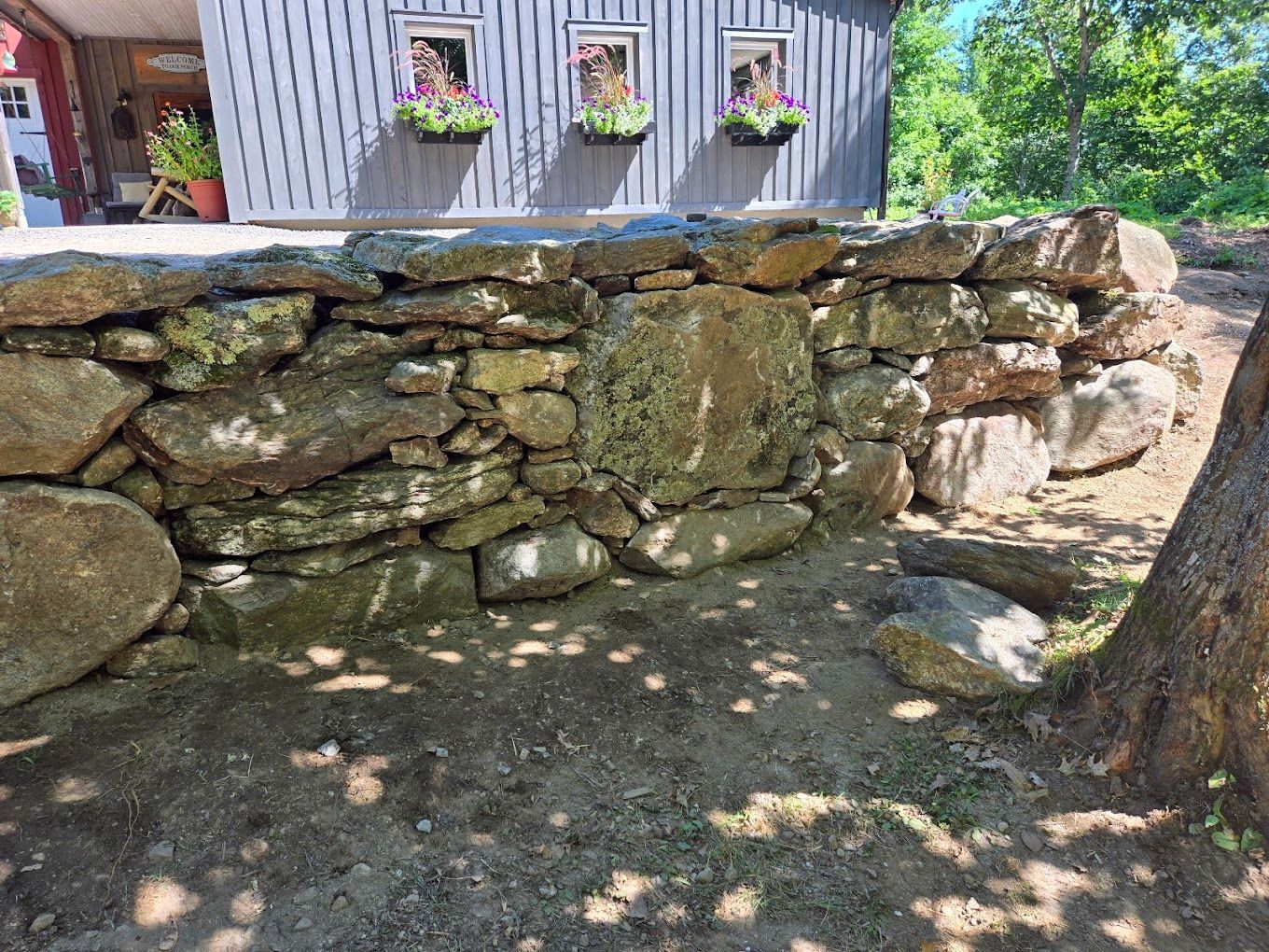 A rustic stone retaining wall sits in front of a blue wooden building with three flower-filled window boxes.