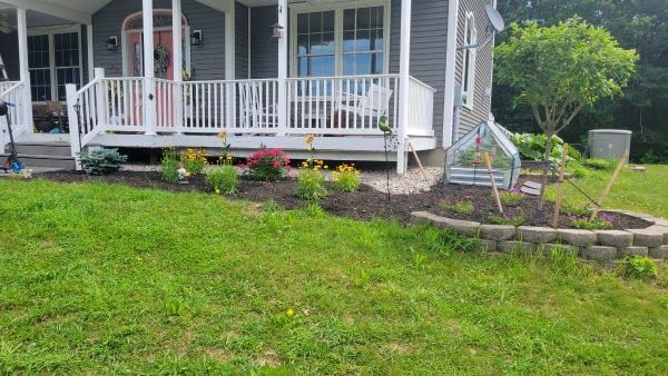 A grey house with a white front porch features a front garden bed with mulch, small green plants, and colorful flowers.