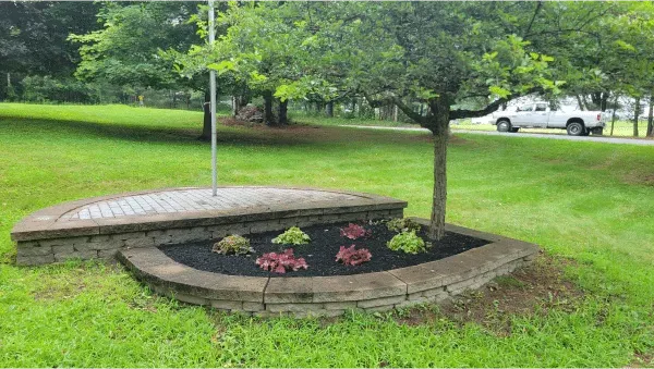 A tiered stone retaining wall garden bed with small plants and mulch sits beneath a tree on a grassy lawn.