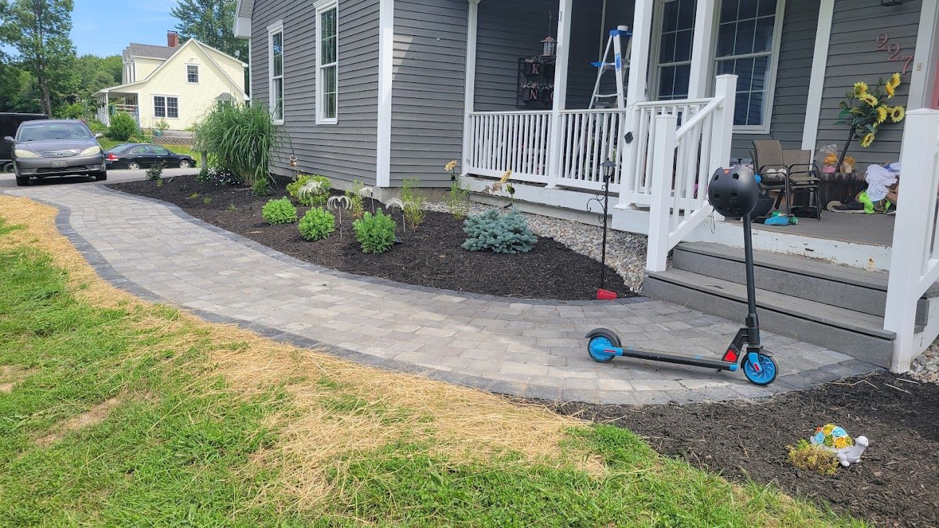 A paver walkway curves along the front of a grey house with a porch, dark mulch, green plants, and a blue scooter.