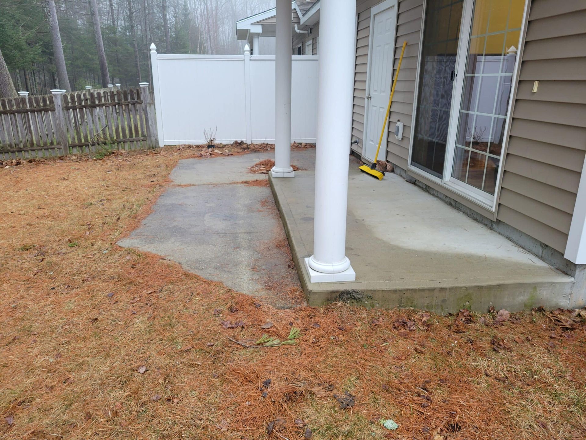 A concrete patio extending from a house with beige siding, white columns, and a white privacy fence in the background.