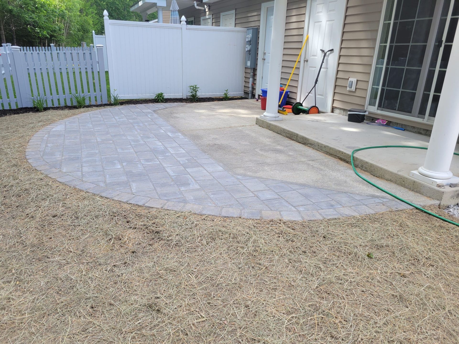 A semi-circular paver patio extension adjacent to a concrete porch and tan siding house, with a white fence in the back.
