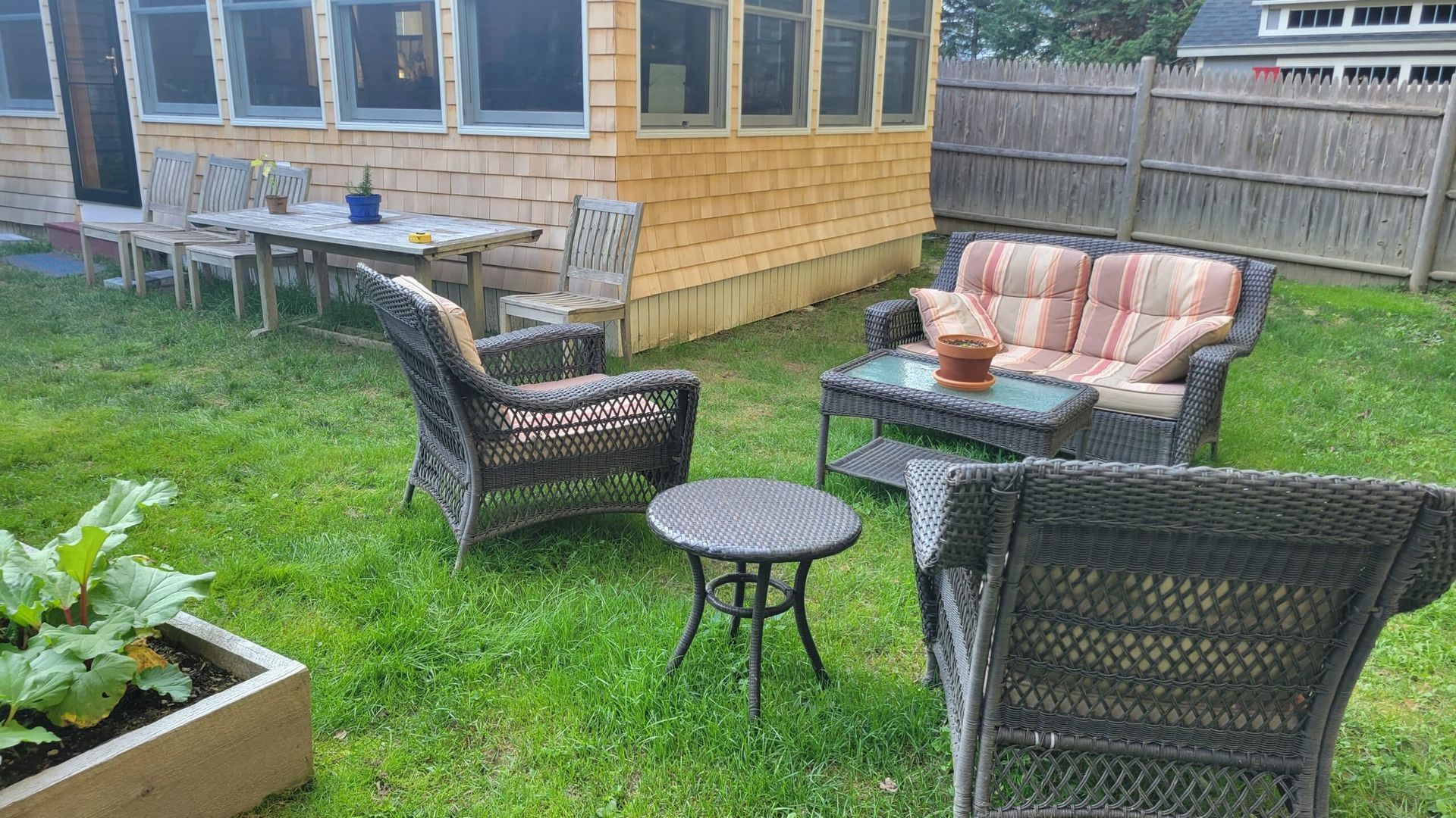 A backyard patio set with wicker chairs, a small table, and a loveseat on the grass near a wooden house and fence.