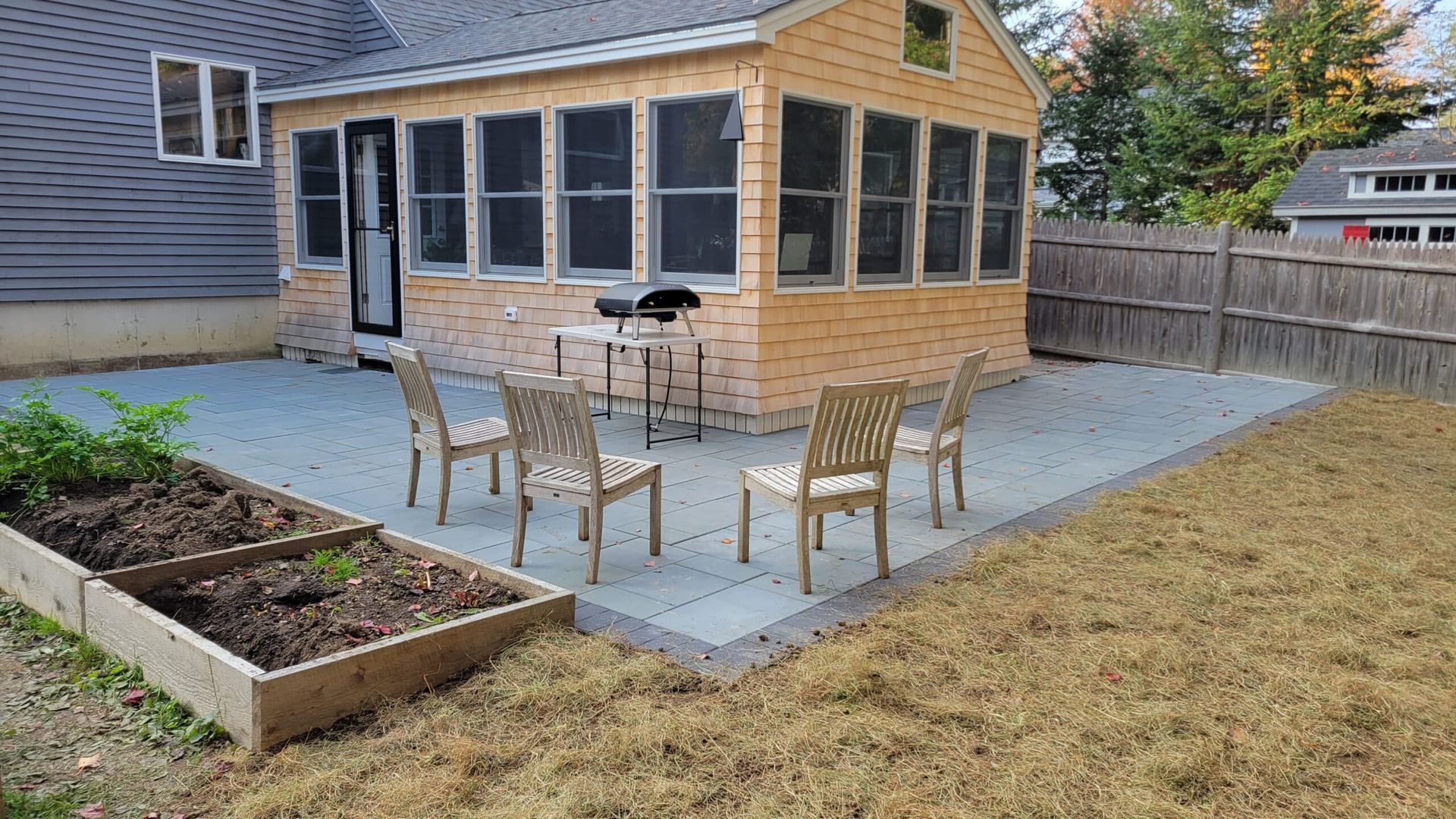 A wooden sunroom with a grey tiled patio, four metal chairs, a small grill, and a raised garden bed in a backyard.