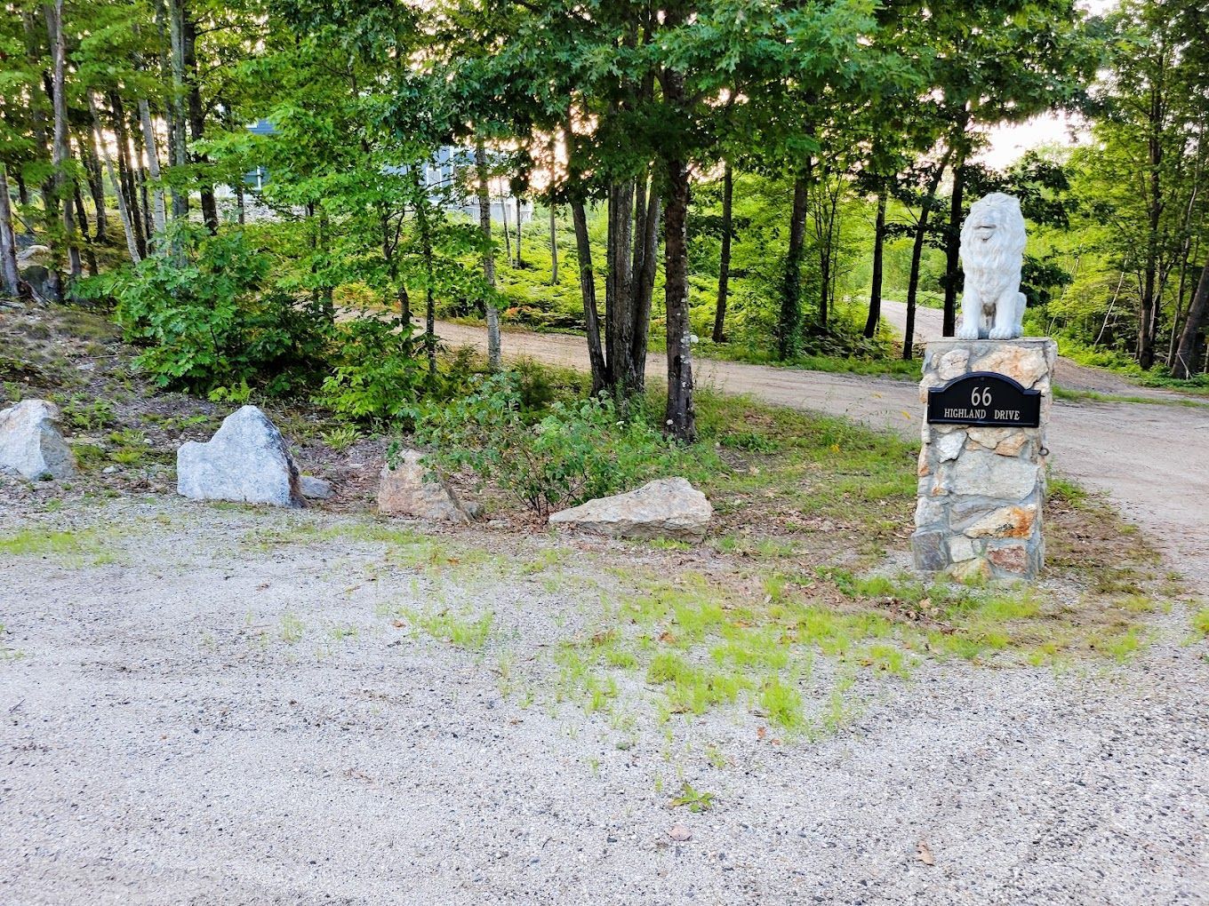 A stone pillar with a white lion statue sits at the entrance of a gravel driveway lined with trees.