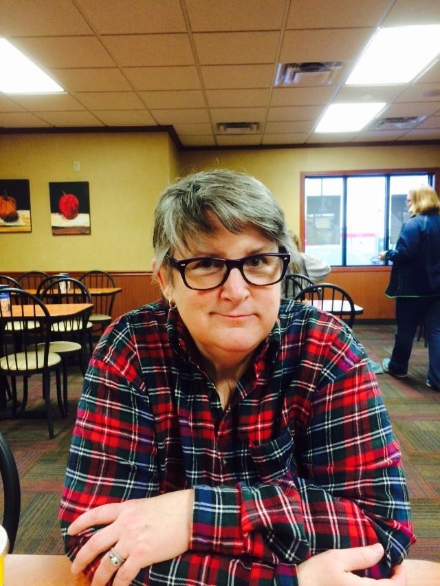 A person with short, gray-brown hair wearing glasses and a red, black, and blue plaid shirt sits at a restaurant table.