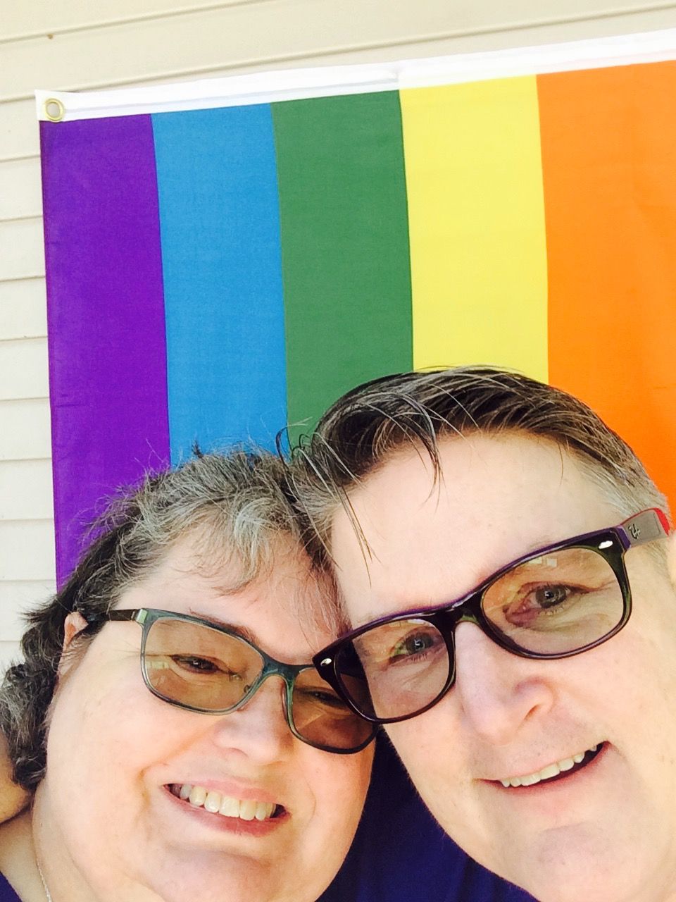Two people wearing glasses smile closely together in front of a rainbow pride flag hanging on a wall.