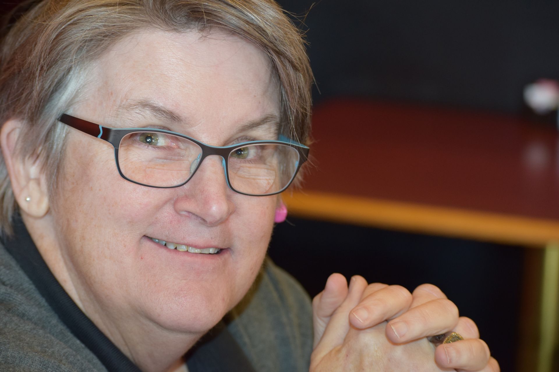 A person with short hair and glasses smiles slightly, hands clasped together in front of them in a professional setting.