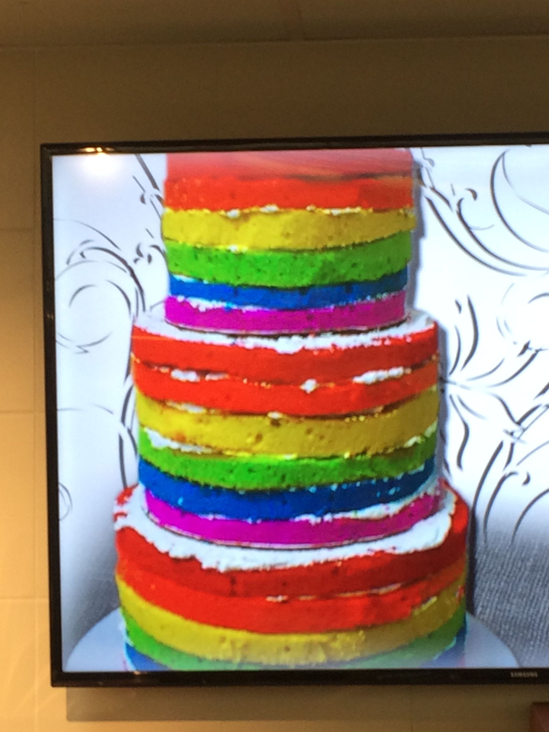 A three-tiered cake with horizontal stripes of red, orange, yellow, green, blue, and purple layers, shown on a screen.