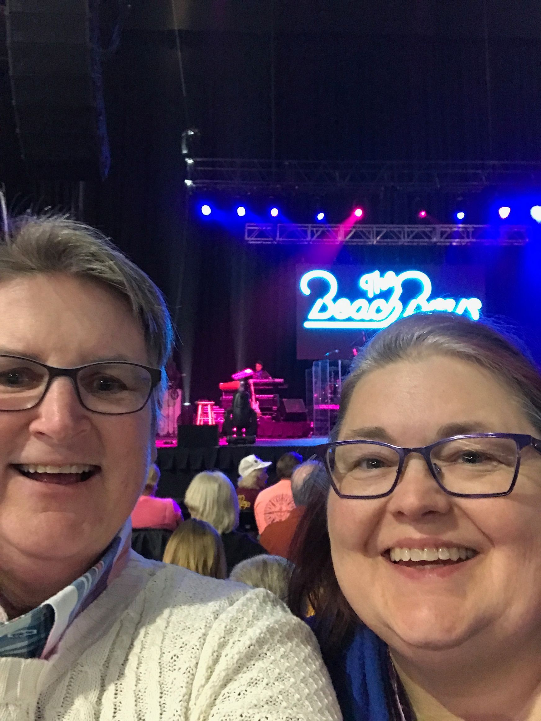 Two smiling people taking a selfie in front of a stage with a bright neon sign reading 