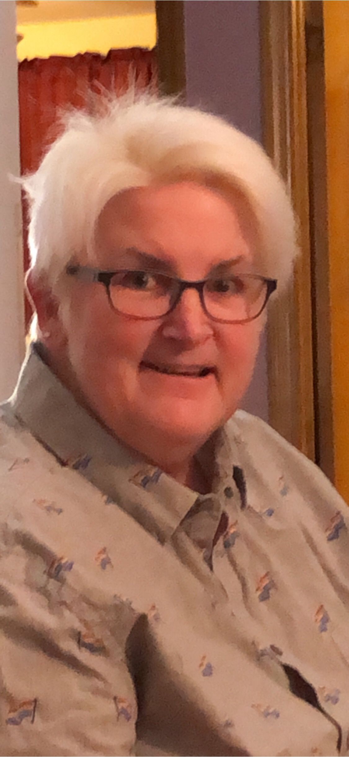 A person with short white hair and glasses smiles while wearing a light-colored button-down shirt with a small pattern.