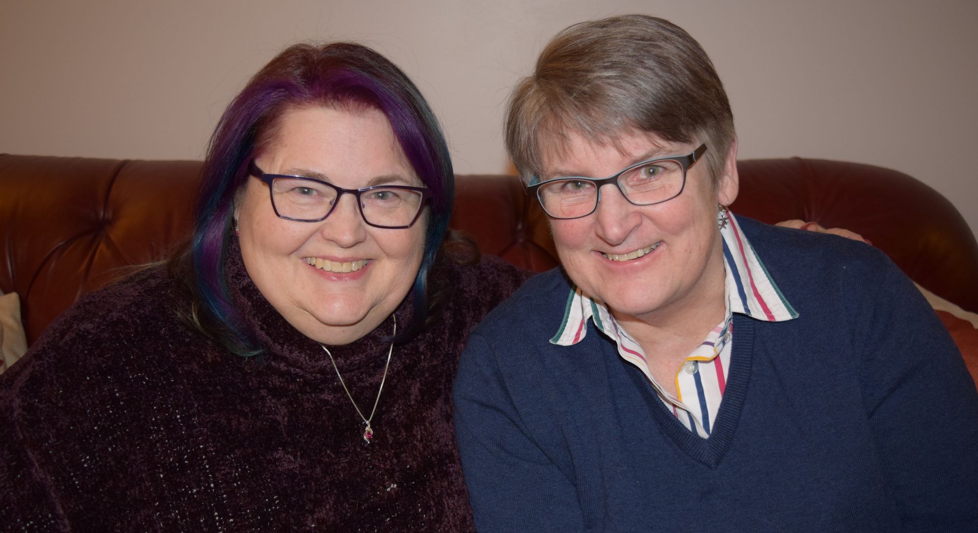 Two people smiling, side-by-side, on a brown leather couch. Both are wearing glasses.