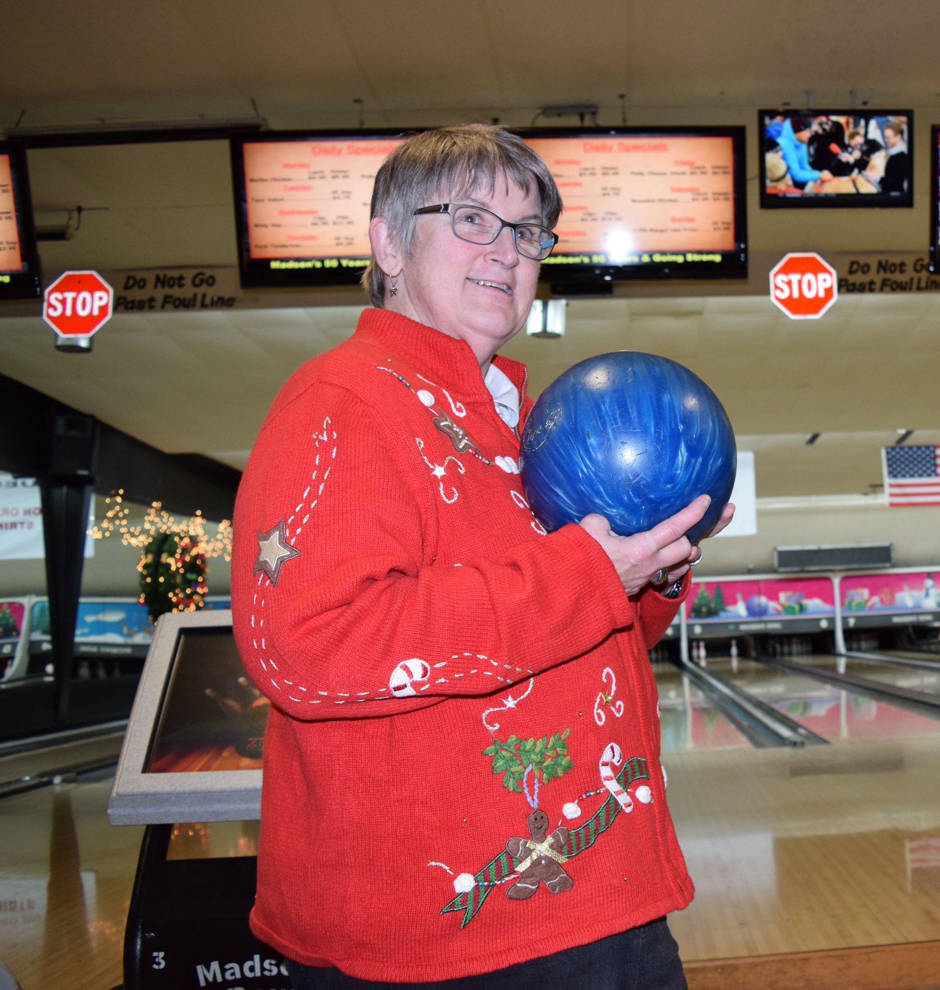 A person wearing a bright red festive sweater holding a blue bowling ball in a bowling alley.