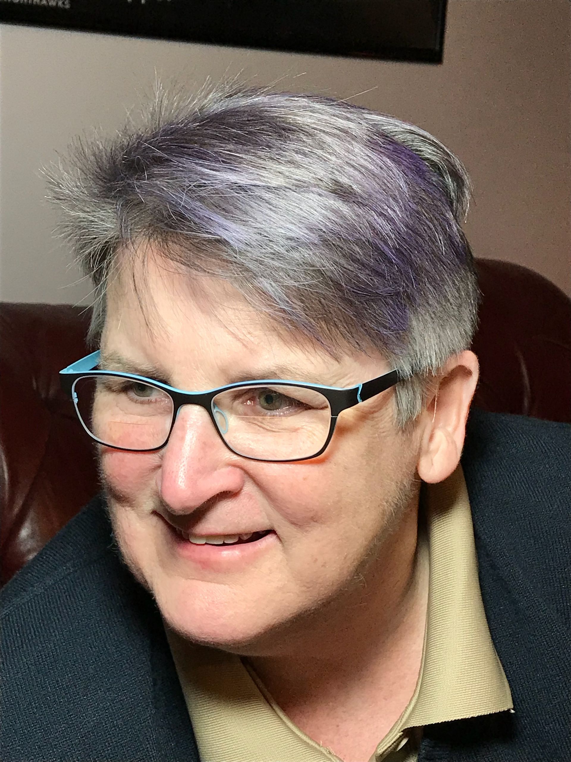 A person wearing glasses with blue and black frames, sporting short, gray-lavender hair, smiling in a dark room.