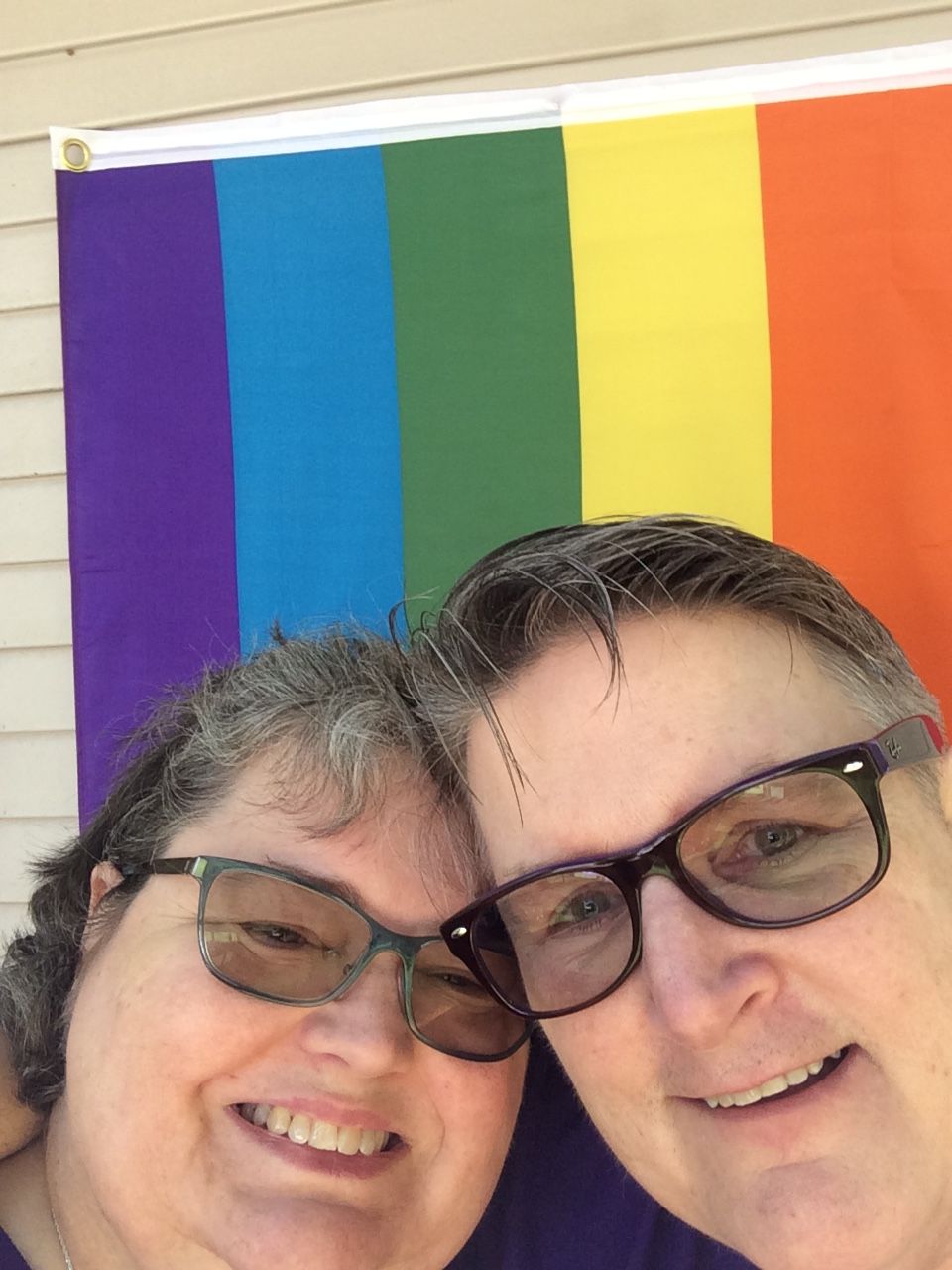 Two smiling people pose together in front of a colorful striped Pride flag.
