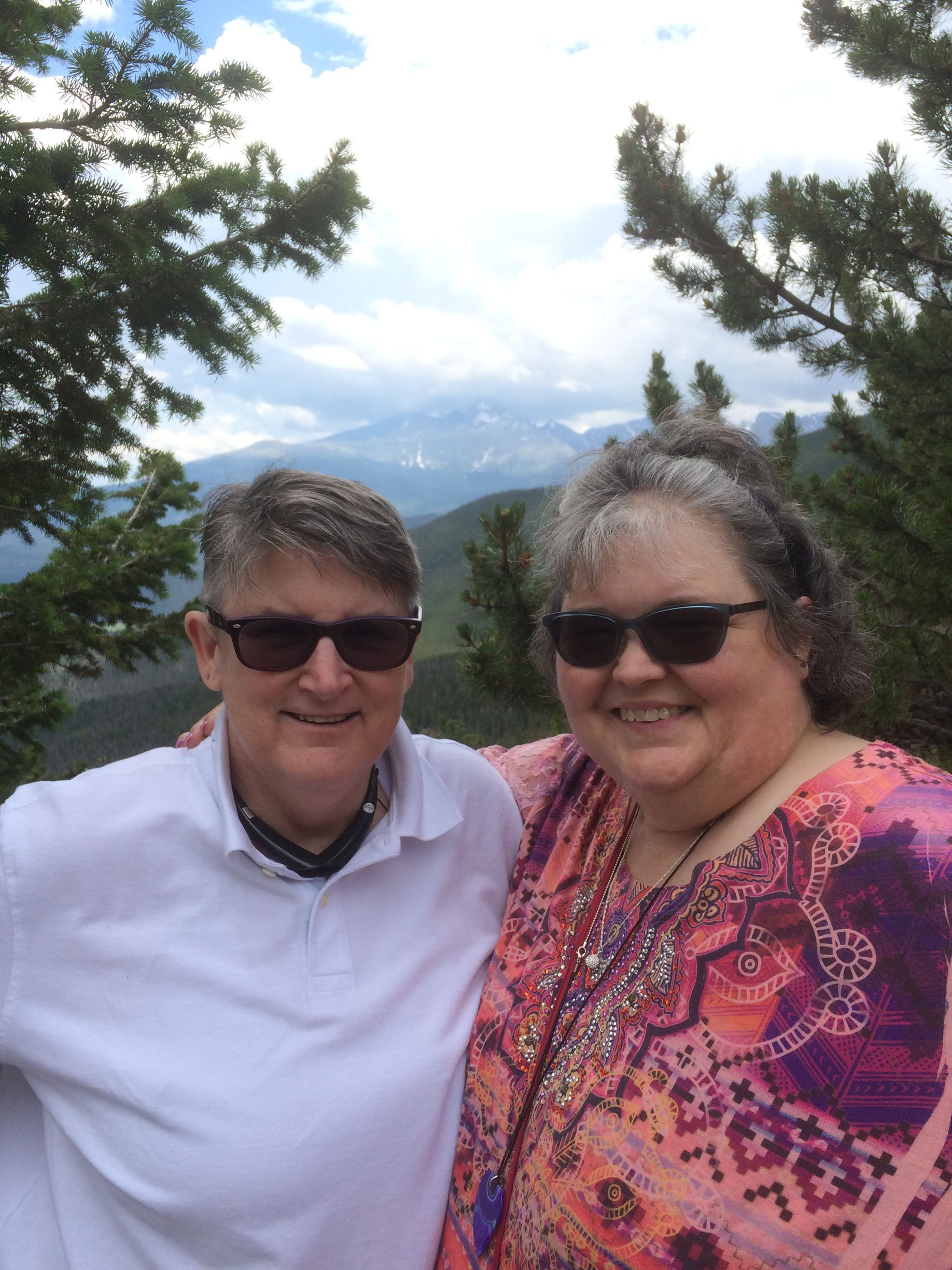 Two people smiling while standing outdoors in front of a mountainous, forest landscape.