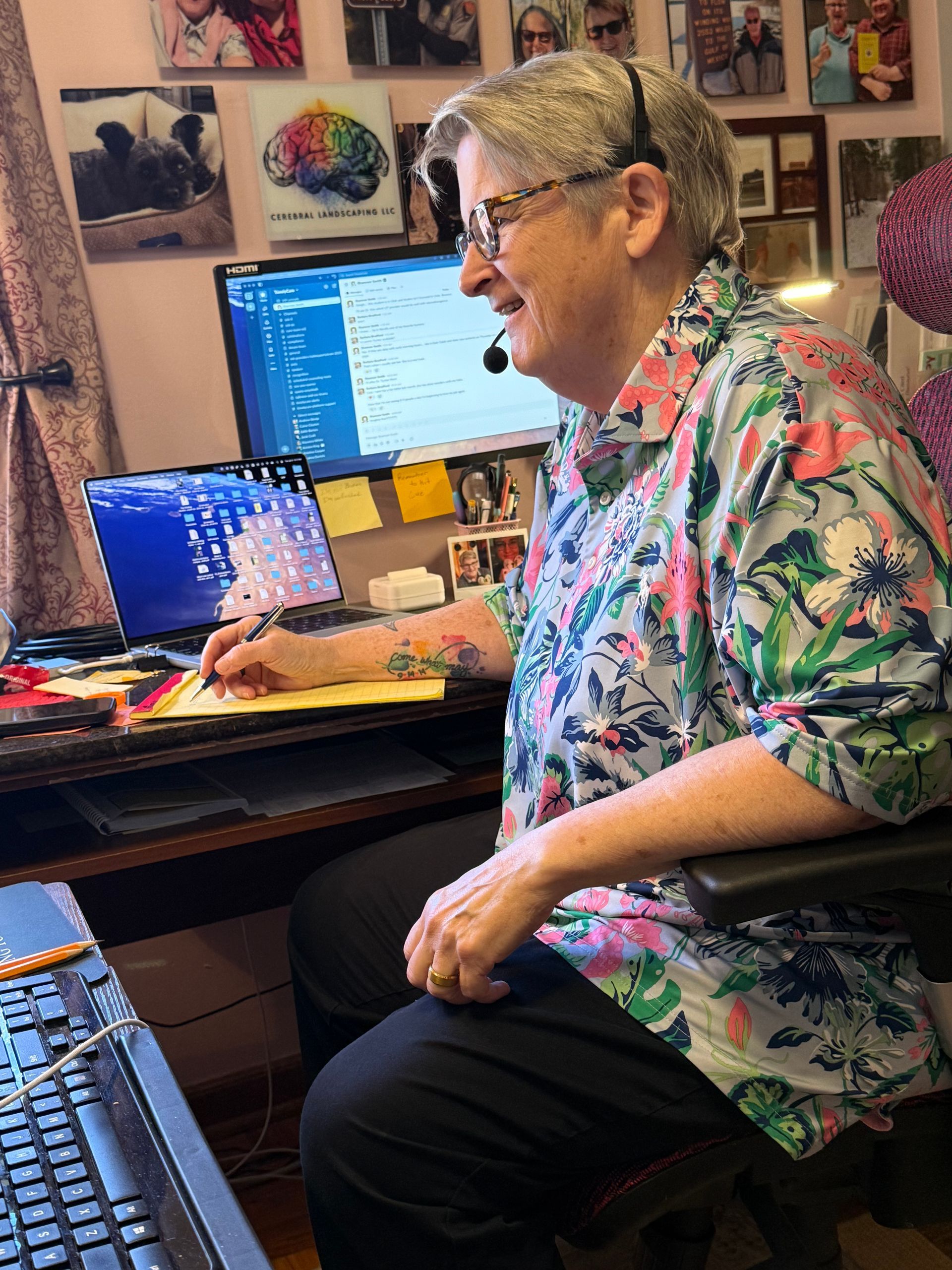 A person wearing a headset and floral shirt writes on a notepad at a desk with two computer screens and wall photos.