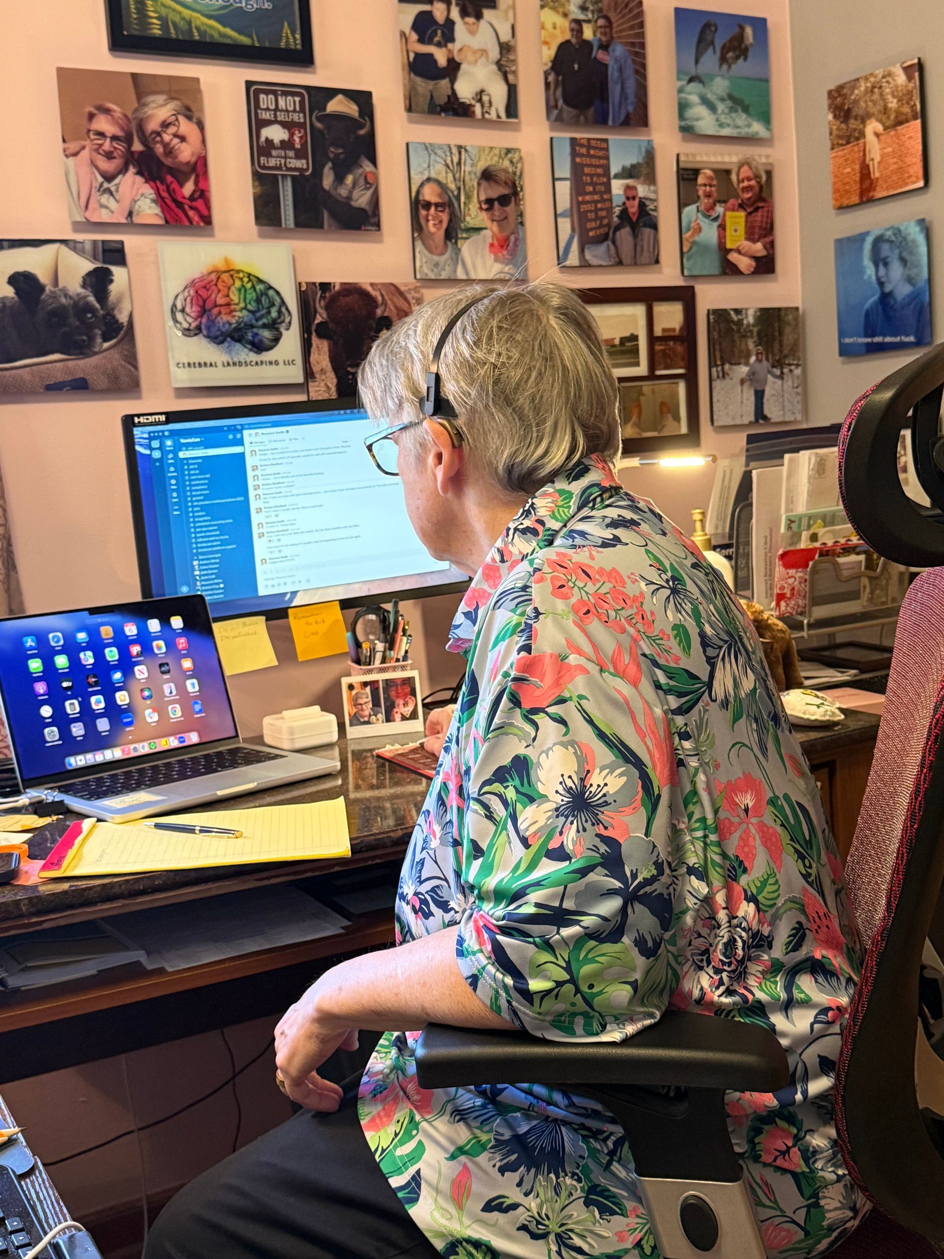 A person wearing a floral shirt sits at a desk with two computer monitors, surrounded by wall-mounted framed photographs.