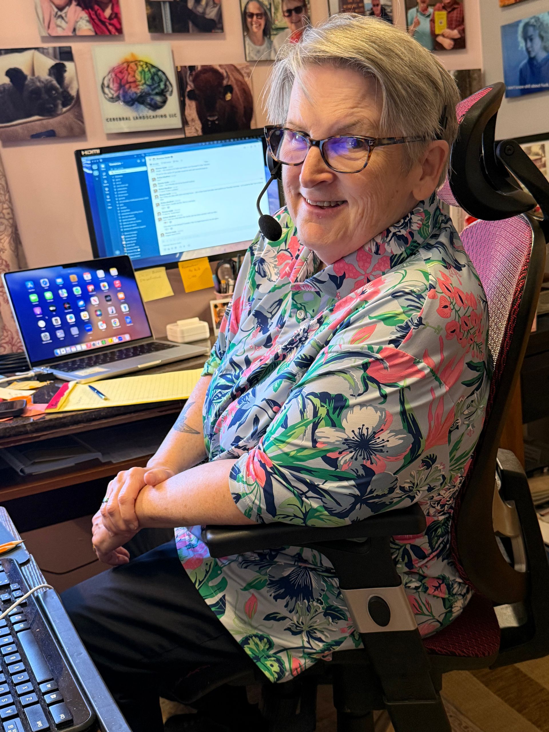 A person with glasses and a headset smiles while seated at a desk with two computer screens in a room with wall decor.