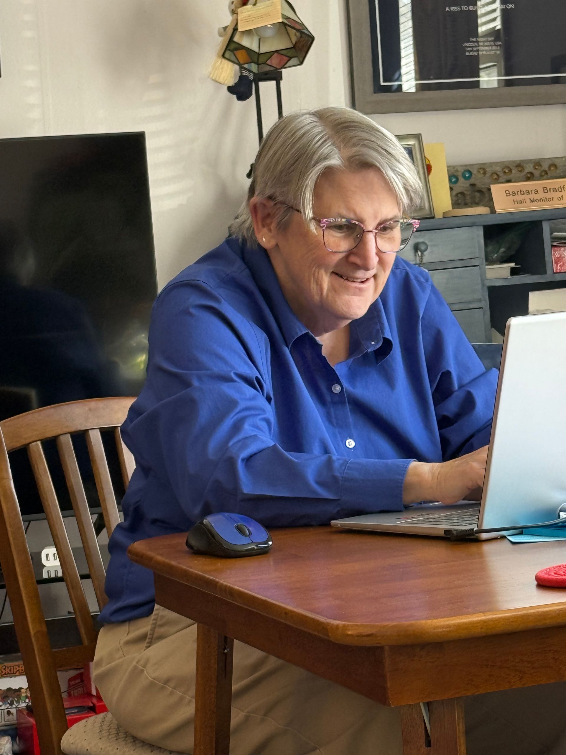 A person wearing a blue collared shirt and glasses sits at a wooden table, smiling while working on a laptop.