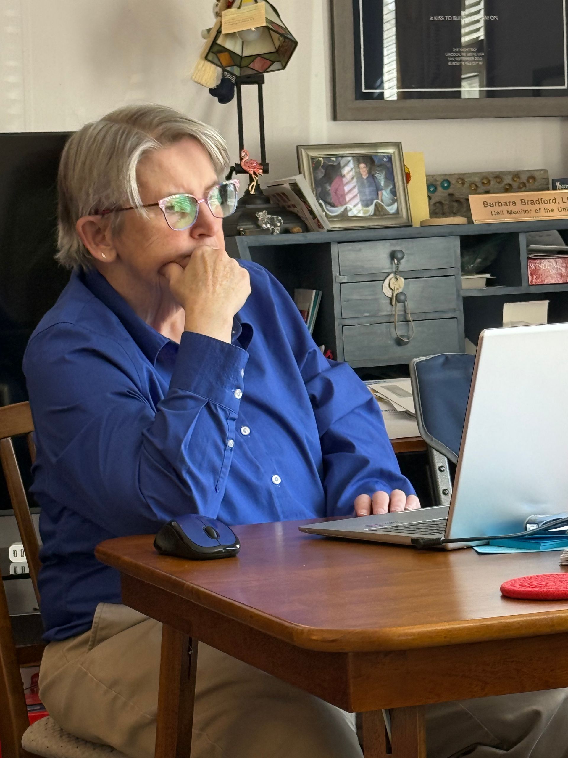 A person in a blue button-down shirt sits at a wooden table, resting their chin on their hand while looking at a laptop.