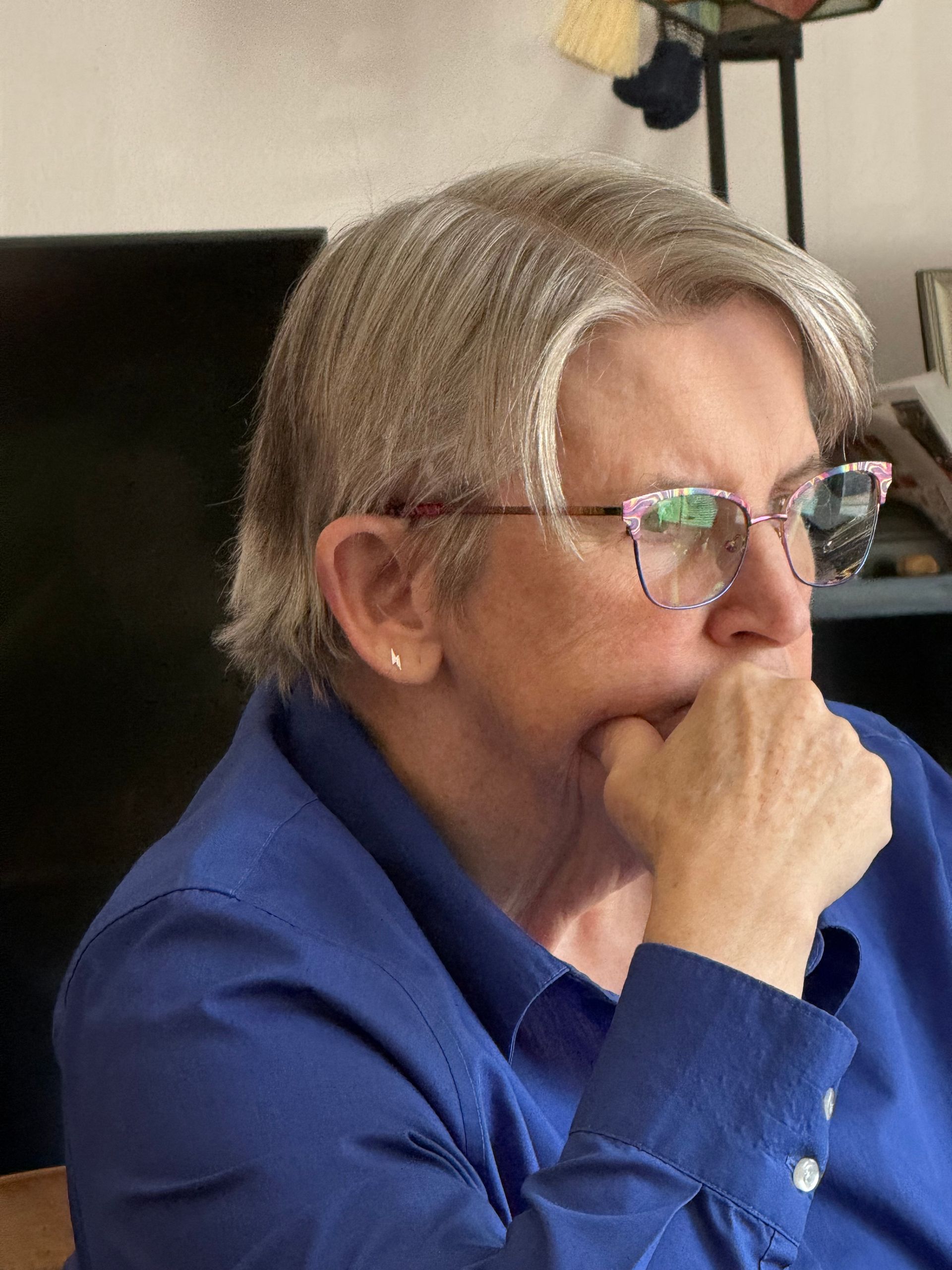 A person with grey hair wearing glasses and a blue button-down shirt, resting their chin on their hand in thought.