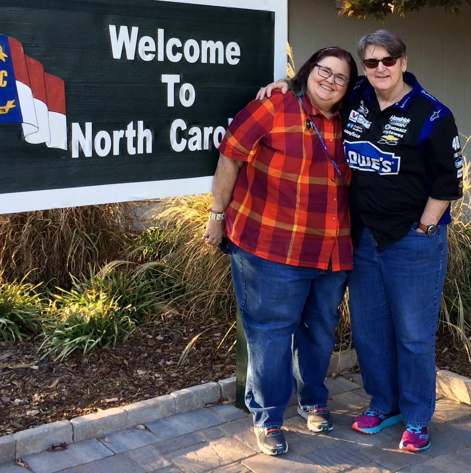 Two people stand side-by-side with arms around each other in front of a 