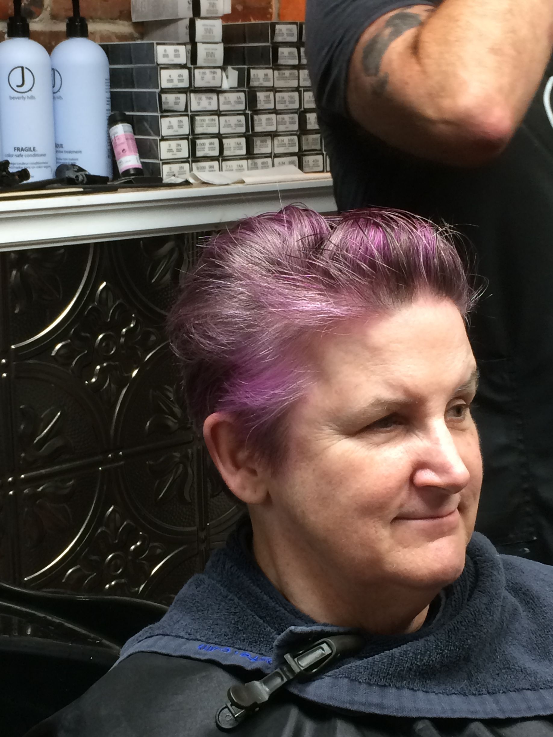 A person with short, vibrant purple hair sits in a salon chair, wearing a protective cape.