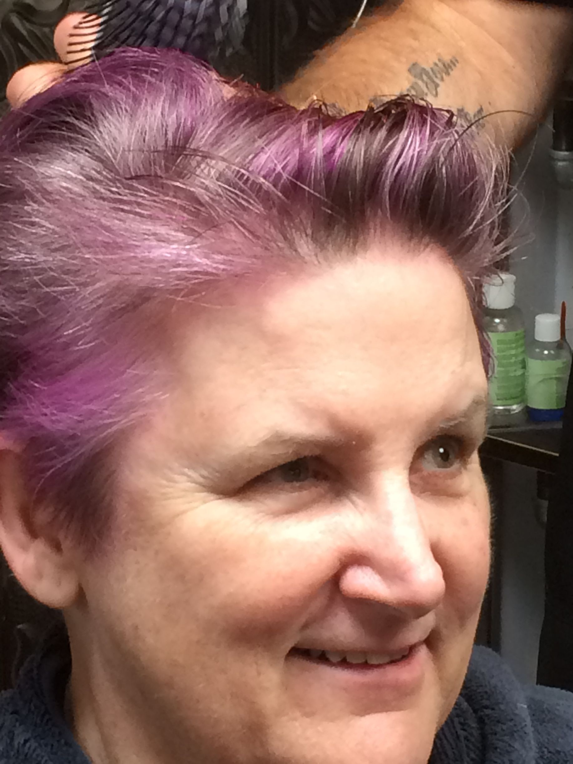 A person with short, vibrant purple hair is smiling while having their hair styled in a salon setting.
