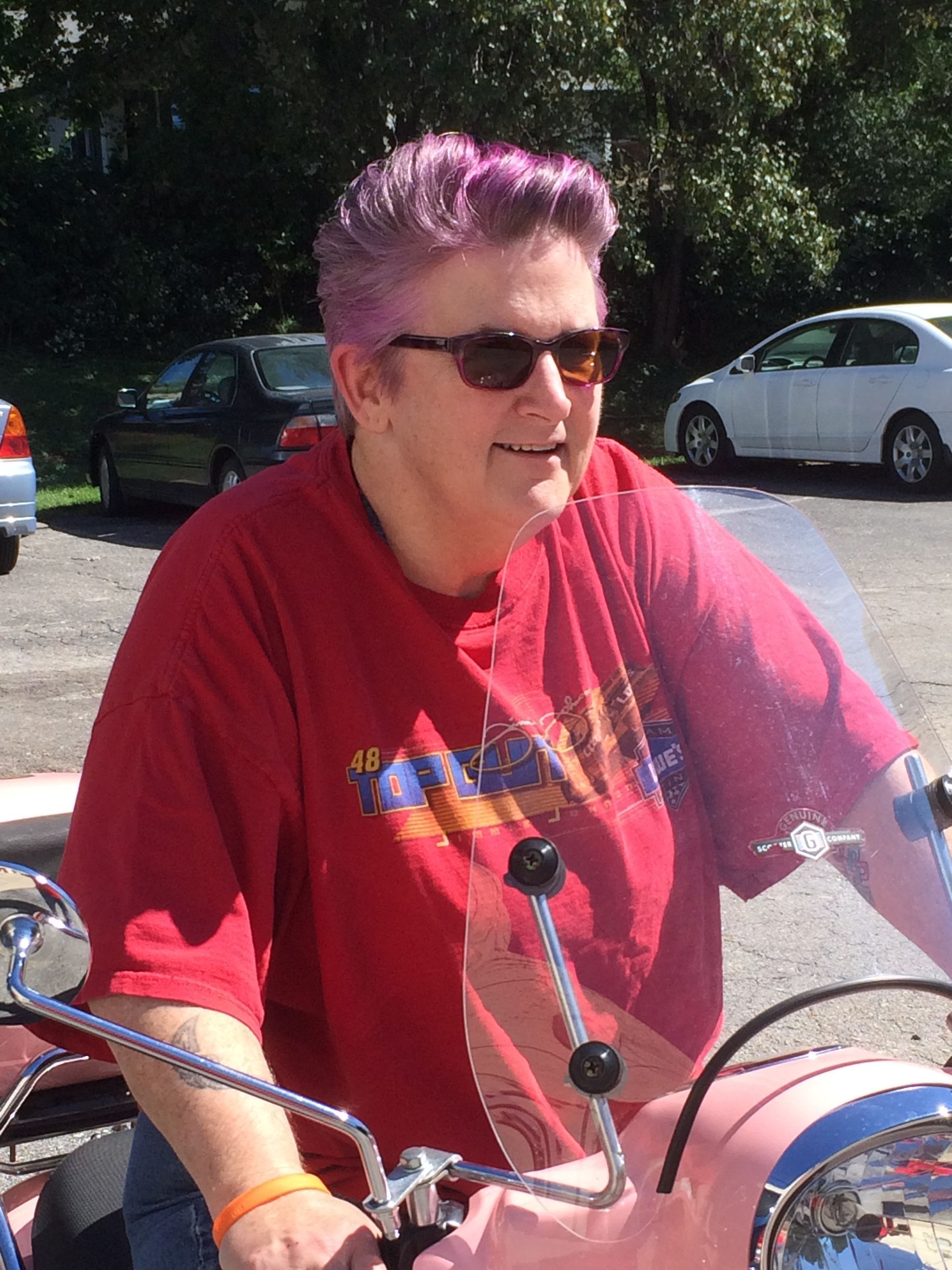 A person with short, magenta hair wearing sunglasses and a red shirt, sitting on a light-pink motorcycle outdoors.