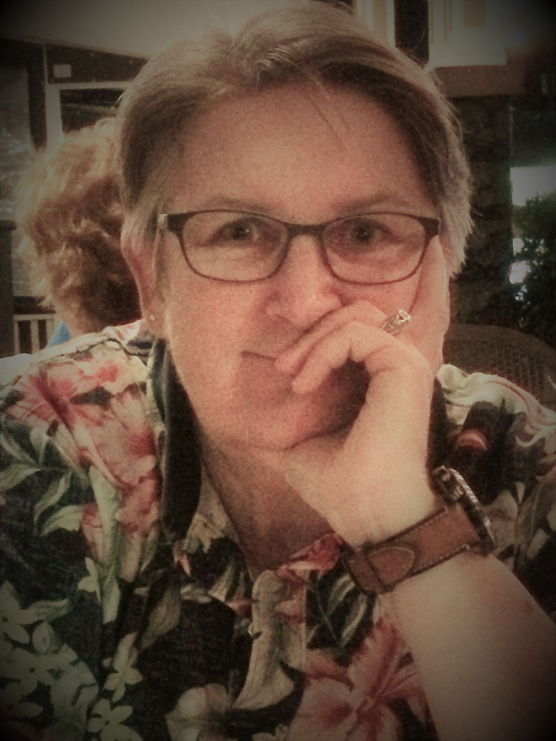 A person with short hair and glasses wearing a floral shirt, resting their chin on their hand while looking at the camera.