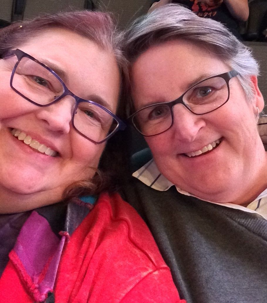 Two people smiling closely together for a selfie, both wearing glasses and casual clothing.