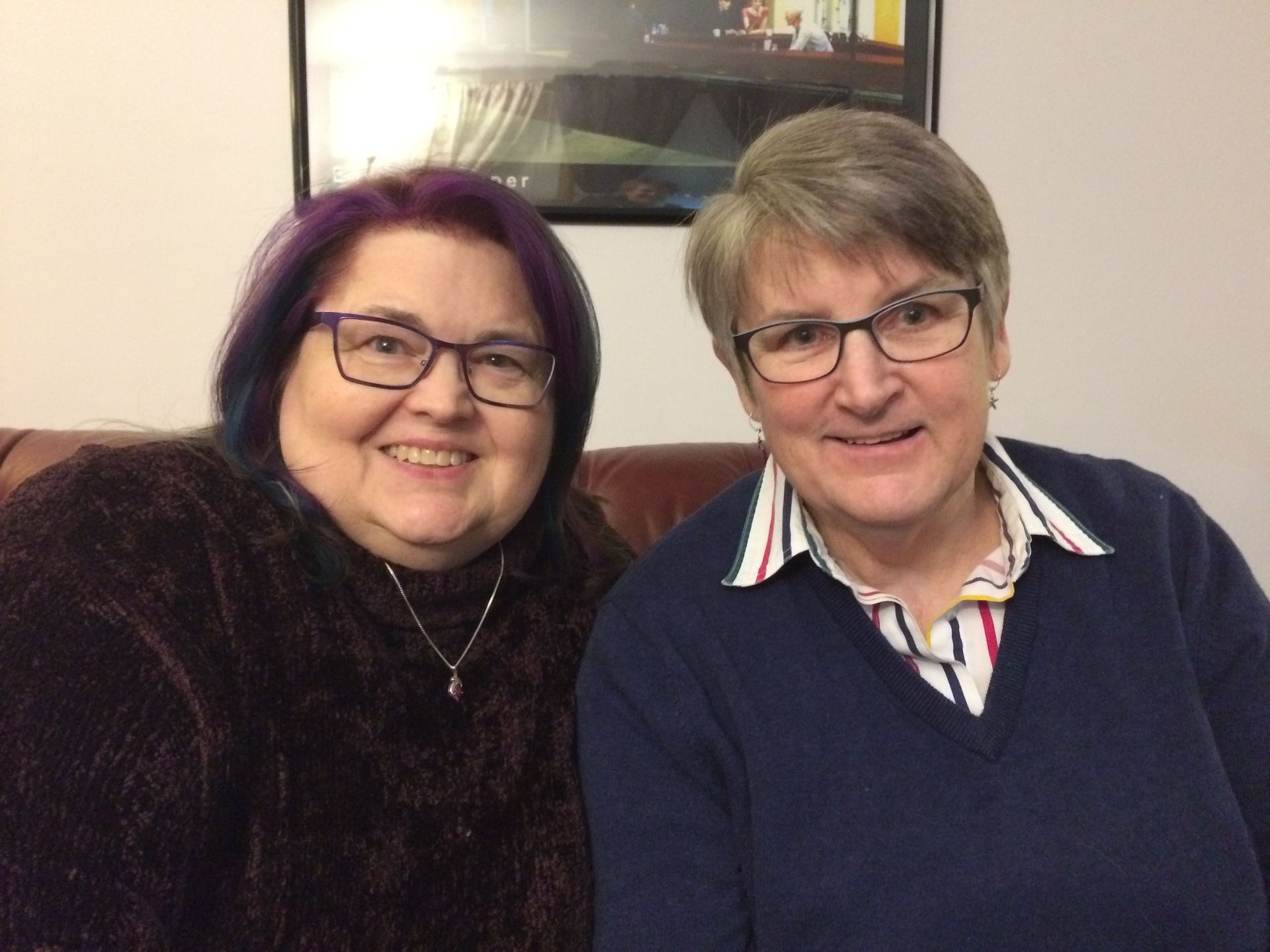 Two people with glasses smiling side-by-side in front of a framed picture on a wall.