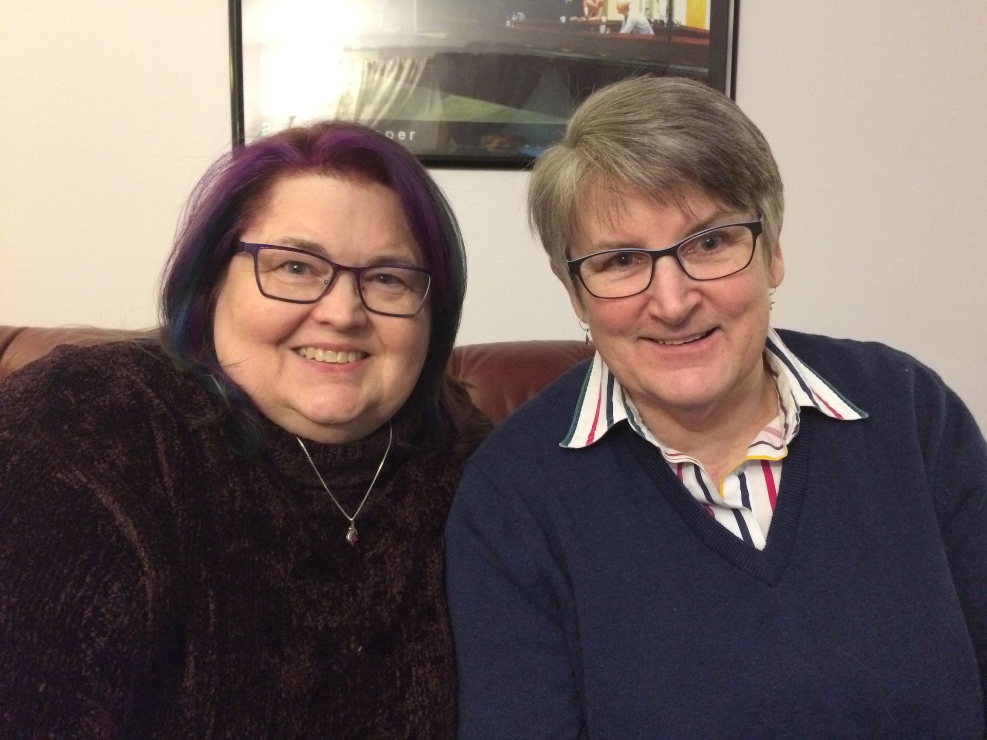 Two people with short hair and glasses smile while sitting side by side on a leather couch in an indoor setting.
